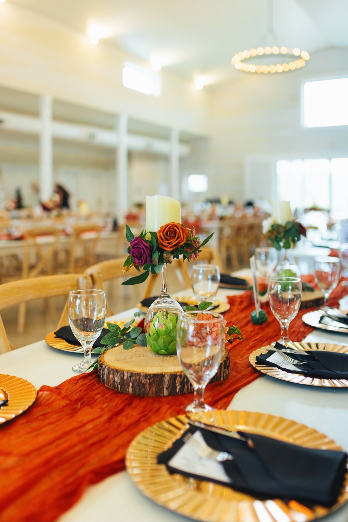 A dressed reception table — gold chargers, rust runner, and a rose-and-candle centerpiece — in a bright venue