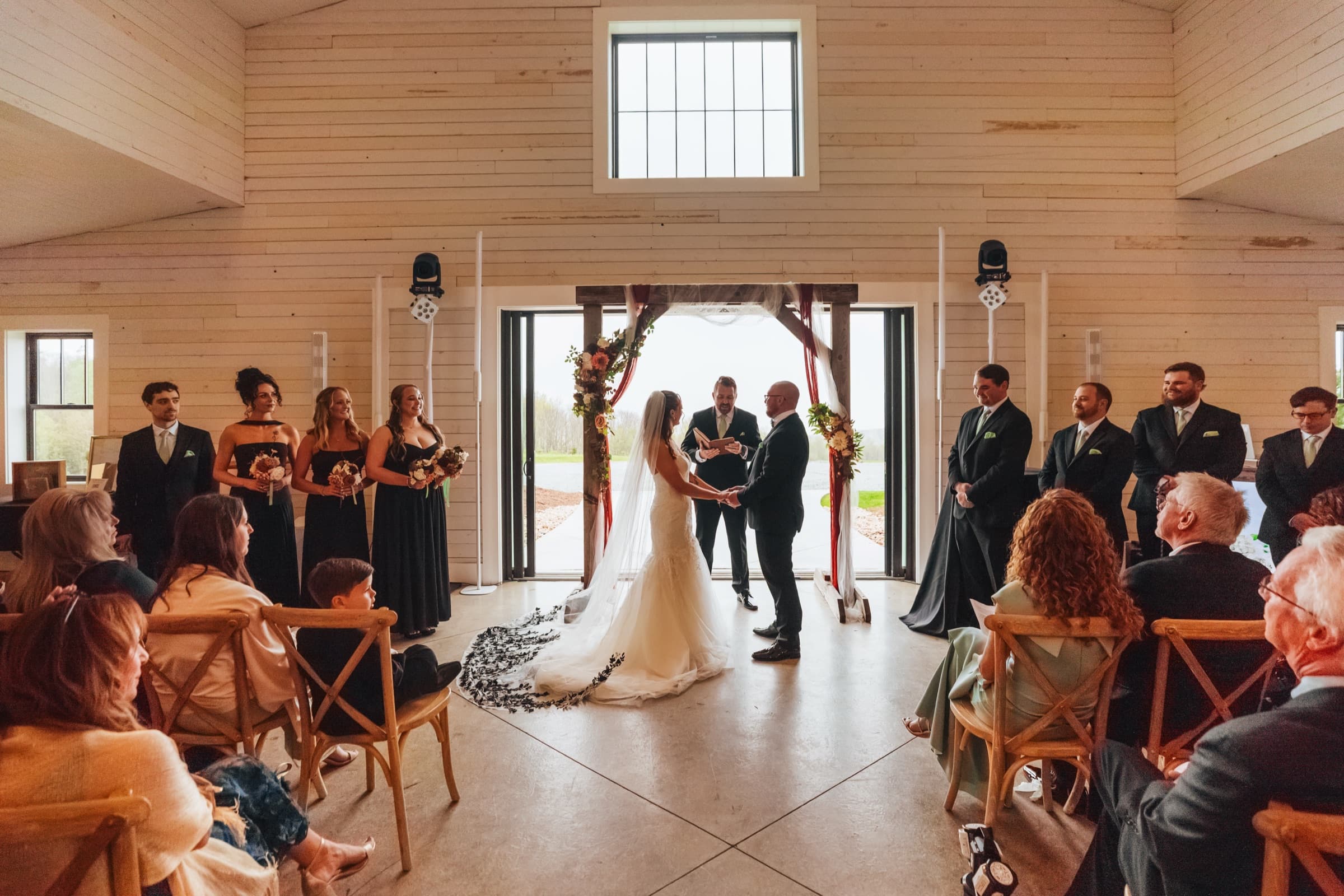 Jami and JJ at the altar inside a white-pine barn, mountains visible through the open doors behind them